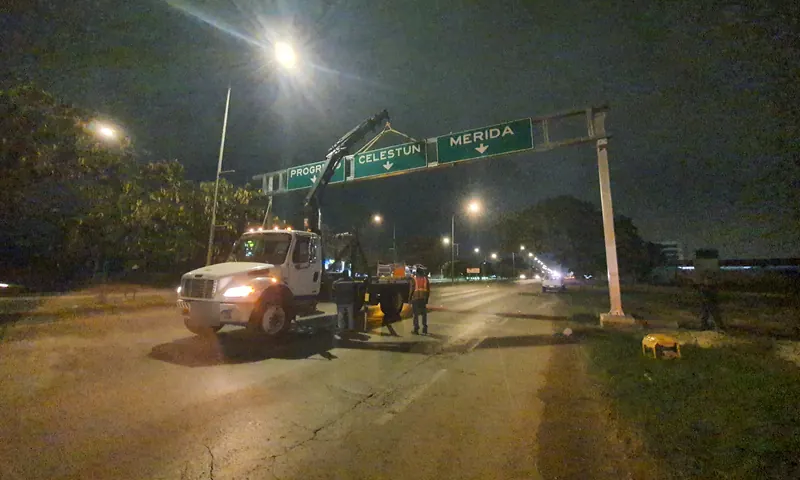 La grúa articulada junto a un marco elevado con los tableros «Progreso», «Celestún» y «Mérida», de noche