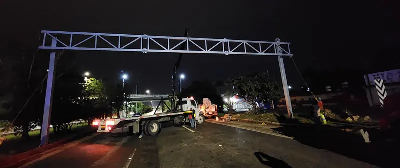 Un marco elevado de acero recién montado atraviesa la vía de noche; debajo, la grúa articulada con su cama plana