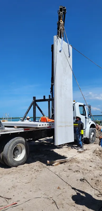 Un panel de concreto de varios metros cuelga vertical del gancho de la grúa articulada, junto al camión, con el mar al fondo